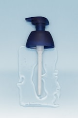 A blue pump dispenser head is lying on a surface with some clear liquid spread around it. The background is a light blue color, providing a clean and minimalistic aesthetic.