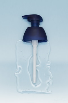 A blue pump dispenser head is lying on a surface with some clear liquid spread around it. The background is a light blue color, providing a clean and minimalistic aesthetic.