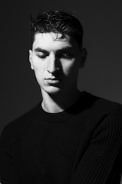 A black and white portrait of a young man with a contemplative expression, wearing a dark sweater. The lighting creates dramatic shadows on his face, emphasizing his features in a minimalist setting.