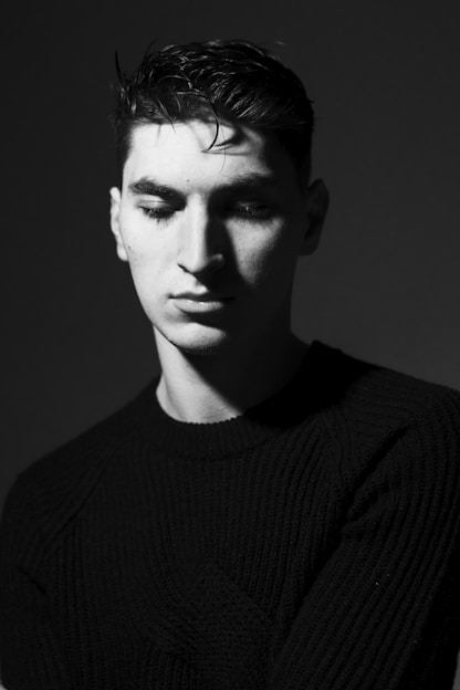 A black and white portrait of a young man with a contemplative expression, wearing a dark sweater. The lighting creates dramatic shadows on his face, emphasizing his features in a minimalist setting.