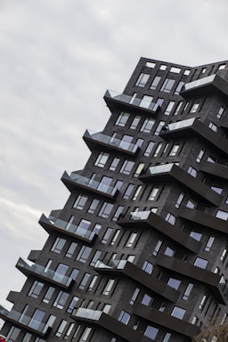 An angular, modern apartment building with dark brick facade and numerous balconies protruding in a stepped formation. The architecture features a unique, staggered design that creates a dynamic visual impact. Large windows cover the facade, reflecting the overcast sky.