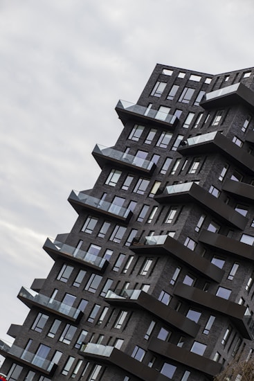 An angular, modern apartment building with dark brick facade and numerous balconies protruding in a stepped formation. The architecture features a unique, staggered design that creates a dynamic visual impact. Large windows cover the facade, reflecting the overcast sky.