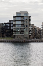 A modern, multi-story apartment building with glass balconies is positioned along a waterfront, with neighboring buildings visible in the background. The scene is overcast, creating a muted, calm atmosphere.