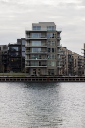 A modern, multi-story apartment building with glass balconies is positioned along a waterfront, with neighboring buildings visible in the background. The scene is overcast, creating a muted, calm atmosphere.