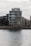 A modern, multi-story apartment building with glass balconies is positioned along a waterfront, with neighboring buildings visible in the background. The scene is overcast, creating a muted, calm atmosphere.