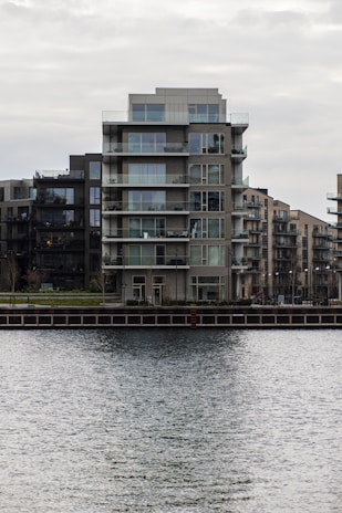 A modern, multi-story apartment building with glass balconies is positioned along a waterfront, with neighboring buildings visible in the background. The scene is overcast, creating a muted, calm atmosphere.