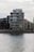 A modern, multi-story apartment building with glass balconies is positioned along a waterfront, with neighboring buildings visible in the background. The scene is overcast, creating a muted, calm atmosphere.