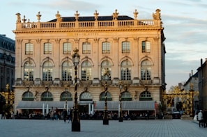 A grand, historic hotel building with ornate architecture and detailed decorations, bathed in warm sunlight. The façade features large arched windows and intricate gold-colored embellishments. There are street lamps and ornamental fencing in front of the building. There are people walking and sitting at outdoor cafes on the street level, contributing to the lively atmosphere.