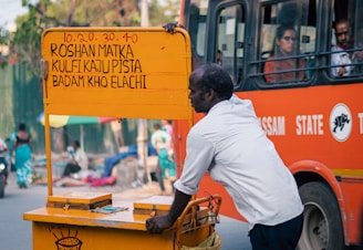 A man in a white shirt pushes a yellow cart with a sign displaying pricing and items like kulfi and pista. In the background, an orange bus carries passengers, and there is pedestrian activity on the street.