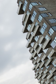 An angular, modern residential building with numerous balconies and large windows. The structure appears tilted, creating a dynamic and somewhat surreal visual effect. The sky is overcast with soft, diffused lighting.