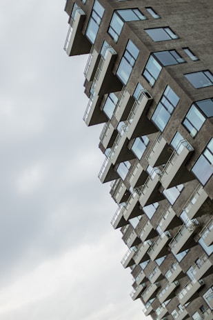 An angular, modern residential building with numerous balconies and large windows. The structure appears tilted, creating a dynamic and somewhat surreal visual effect. The sky is overcast with soft, diffused lighting.