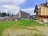 A resort area with grassy fields in the foreground, featuring a large ski lift structure on the left. Buildings, including a tall hotel and a traditional chalet-style lodge, are visible. Pine trees border the scene, under a partly cloudy sky.