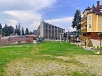 A resort area with grassy fields in the foreground, featuring a large ski lift structure on the left. Buildings, including a tall hotel and a traditional chalet-style lodge, are visible. Pine trees border the scene, under a partly cloudy sky.