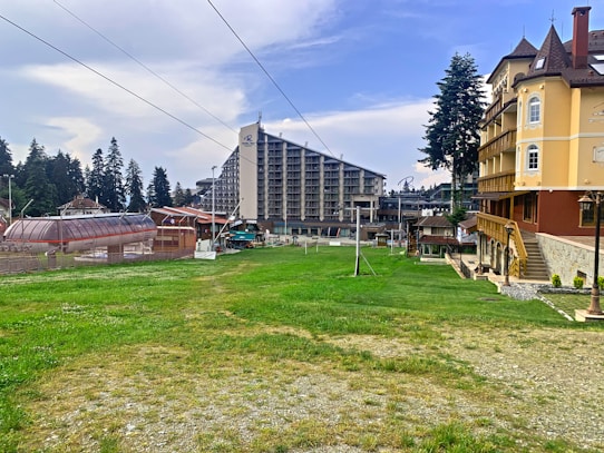 A resort area with grassy fields in the foreground, featuring a large ski lift structure on the left. Buildings, including a tall hotel and a traditional chalet-style lodge, are visible. Pine trees border the scene, under a partly cloudy sky.