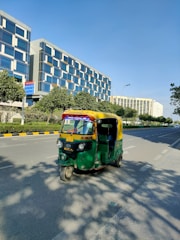 An auto-rickshaw is driving on a wide, empty road flanked by modern office buildings and green trees. The auto-rickshaw is painted in bright green and yellow colors. It's sunny with a clear blue sky. The buildings have reflective glass windows and appear tall and contemporary in design. The road has marked lanes with sidewalks and some shadows of the trees visible.