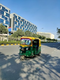 An auto-rickshaw is driving on a wide, empty road flanked by modern office buildings and green trees. The auto-rickshaw is painted in bright green and yellow colors. It's sunny with a clear blue sky. The buildings have reflective glass windows and appear tall and contemporary in design. The road has marked lanes with sidewalks and some shadows of the trees visible.