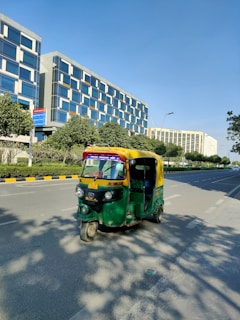 An auto-rickshaw is driving on a wide, empty road flanked by modern office buildings and green trees. The auto-rickshaw is painted in bright green and yellow colors. It's sunny with a clear blue sky. The buildings have reflective glass windows and appear tall and contemporary in design. The road has marked lanes with sidewalks and some shadows of the trees visible.