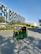 An auto-rickshaw is driving on a wide, empty road flanked by modern office buildings and green trees. The auto-rickshaw is painted in bright green and yellow colors. It's sunny with a clear blue sky. The buildings have reflective glass windows and appear tall and contemporary in design. The road has marked lanes with sidewalks and some shadows of the trees visible.