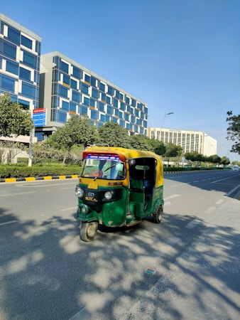 An auto-rickshaw is driving on a wide, empty road flanked by modern office buildings and green trees. The auto-rickshaw is painted in bright green and yellow colors. It's sunny with a clear blue sky. The buildings have reflective glass windows and appear tall and contemporary in design. The road has marked lanes with sidewalks and some shadows of the trees visible.