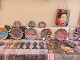 A collection of vibrant decorative plates and artifacts displayed on a patterned fabric. The plates feature intricate designs including fish motifs, floral patterns, and abstract art. A bag with a colorful portrait of a woman hangs above the plates.