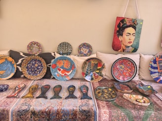 A collection of vibrant decorative plates and artifacts displayed on a patterned fabric. The plates feature intricate designs including fish motifs, floral patterns, and abstract art. A bag with a colorful portrait of a woman hangs above the plates.