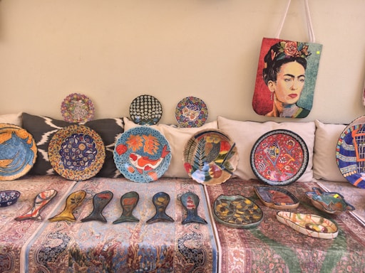A collection of vibrant decorative plates and artifacts displayed on a patterned fabric. The plates feature intricate designs including fish motifs, floral patterns, and abstract art. A bag with a colorful portrait of a woman hangs above the plates.