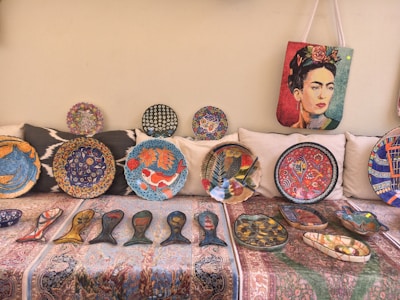 A collection of vibrant decorative plates and artifacts displayed on a patterned fabric. The plates feature intricate designs including fish motifs, floral patterns, and abstract art. A bag with a colorful portrait of a woman hangs above the plates.