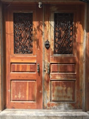 A set of two rustic wooden doors with metal decorative grills in the upper panels. The wood shows signs of weathering and age, with variations in color and texture. Each door has an ornate metal handle, and a knocker is present on the right door. The lighting casts shadows, adding depth to the scene.