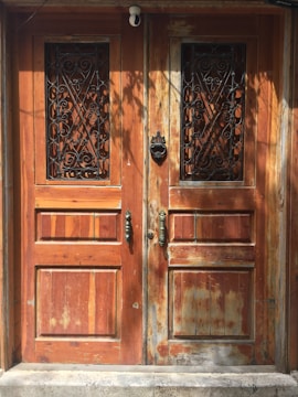 A set of two rustic wooden doors with metal decorative grills in the upper panels. The wood shows signs of weathering and age, with variations in color and texture. Each door has an ornate metal handle, and a knocker is present on the right door. The lighting casts shadows, adding depth to the scene.