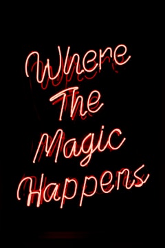 Neon text sign displaying the words Where The Magic Happens in bright red script against a dark background.