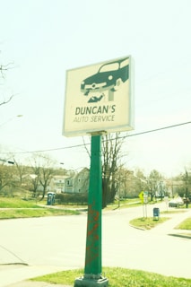 A green pole with a sign for 'Duncan's Auto Service' featuring a graphic of a car being worked on. The background includes a suburban street scene with houses, trees, a mailbox, and a traffic sign. The overall lighting suggests a bright, sunny day.