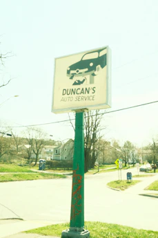 A green pole with a sign for 'Duncan's Auto Service' featuring a graphic of a car being worked on. The background includes a suburban street scene with houses, trees, a mailbox, and a traffic sign. The overall lighting suggests a bright, sunny day.