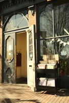 A quaint storefront features a vintage-style door and a large window. Beside the door, there is a cart displaying vinyl records. The brick exterior and decorative wrought iron elements add to the charming aesthetic. Sunlight casts warm tones on the scene, and a small sign with a logo is visible on the door.
