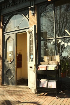 A quaint storefront features a vintage-style door and a large window. Beside the door, there is a cart displaying vinyl records. The brick exterior and decorative wrought iron elements add to the charming aesthetic. Sunlight casts warm tones on the scene, and a small sign with a logo is visible on the door.