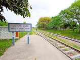 A railway station sign is prominently displayed on the left, with text in both Urdu and English indicating Margalla Railway Station, Islamabad. The sign is set against a backdrop of lush green trees and foliage. Two railway tracks run parallel to each other, leading into the distance. A person can be seen walking on the platform next to the tracks. The ground is paved, and there are leaves scattered on the platform.