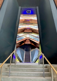 A modern architectural structure featuring a staircase with metal railings leading upwards, framed by geometric patterns. The design is visually striking with contrasting dark and metallic tones, and a neon-like blue light accent at the top.