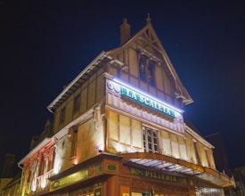 A historic-looking multi-story building with Tudor-style architecture, featuring illuminated signage reading 'La Scaleta' and 'Ristorante Pizzeria'. The structure is adorned with wooden beams, and the glow from the signage offers a warm, inviting ambiance against a dark night sky.