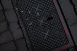 A dark, wet street surface with a textured drainage cover featuring a grid-like pattern and some red markings. The surrounding area is paved with cobblestones.