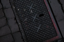 A dark, wet street surface with a textured drainage cover featuring a grid-like pattern and some red markings. The surrounding area is paved with cobblestones.