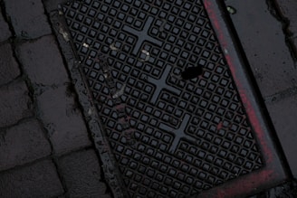 A dark, wet street surface with a textured drainage cover featuring a grid-like pattern and some red markings. The surrounding area is paved with cobblestones.