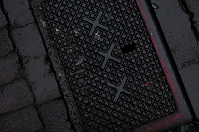 A dark, wet street surface with a textured drainage cover featuring a grid-like pattern and some red markings. The surrounding area is paved with cobblestones.