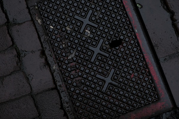A dark, wet street surface with a textured drainage cover featuring a grid-like pattern and some red markings. The surrounding area is paved with cobblestones.