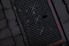 A dark, wet street surface with a textured drainage cover featuring a grid-like pattern and some red markings. The surrounding area is paved with cobblestones.