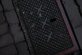 A dark, wet street surface with a textured drainage cover featuring a grid-like pattern and some red markings. The surrounding area is paved with cobblestones.