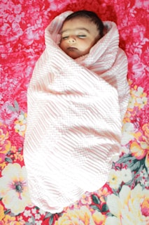 A sleeping baby wrapped in a textured white blanket lies on a vibrant, floral-patterned red and pink background.