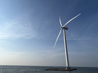 A large wind turbine stands on a small rocky platform in the sea. The sky is clear with some light clouds, conveying a calm atmosphere. The horizon is visible where the sea meets the sky.