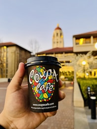 A hand is holding a colorful coffee cup with the text 'Coupa Cafe' printed on it. In the background, there is a large building with a prominent red-roofed tower, and the ambient lighting suggests early evening or morning.
