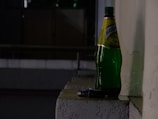 A large green bottle labeled in Cyrillic script is placed on a ledge against a textured wall. Next to the bottle, there is a small package, possibly a cigarette box. The setting appears to be dimly lit, suggestive of evening or nighttime, with shadows cast against the wall.