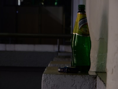 A large green bottle labeled in Cyrillic script is placed on a ledge against a textured wall. Next to the bottle, there is a small package, possibly a cigarette box. The setting appears to be dimly lit, suggestive of evening or nighttime, with shadows cast against the wall.
