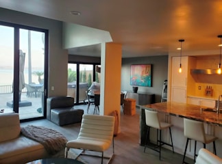 A modern living space with an open-plan design featuring a kitchen, living room, and a view of an outdoor patio through large glass doors. The area is decorated with contemporary furniture, including white chairs and a marble countertop. A colorful abstract painting adorns one wall, and the lighting is warm with hanging ceiling lights.